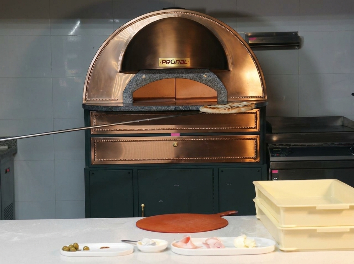 A beautifully baked pizza with a blistered crust being lifted out of a copper oven on a metal pizza peel.
