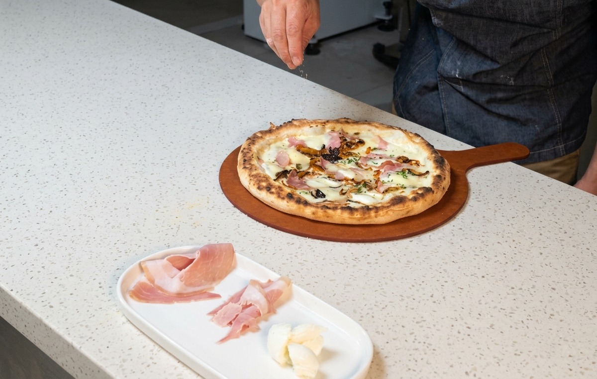 A chefs hand sprinkling dry grated cheese over a hot, freshly baked ham and mushroom pizza.
