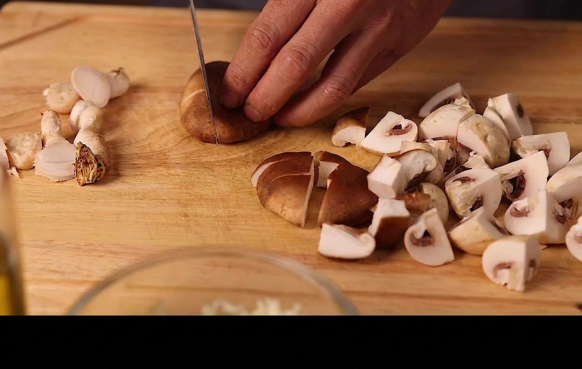 Hands slicing fresh shiitake and button mushrooms into pieces on a wooden cutting board.