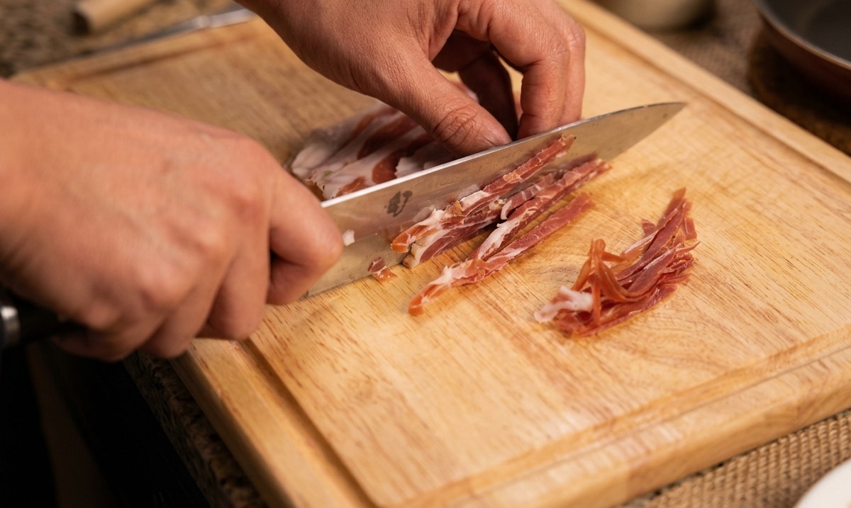 Hands using a chefs knife to cut thin strips of dry-cured ham on a wooden cutting board.