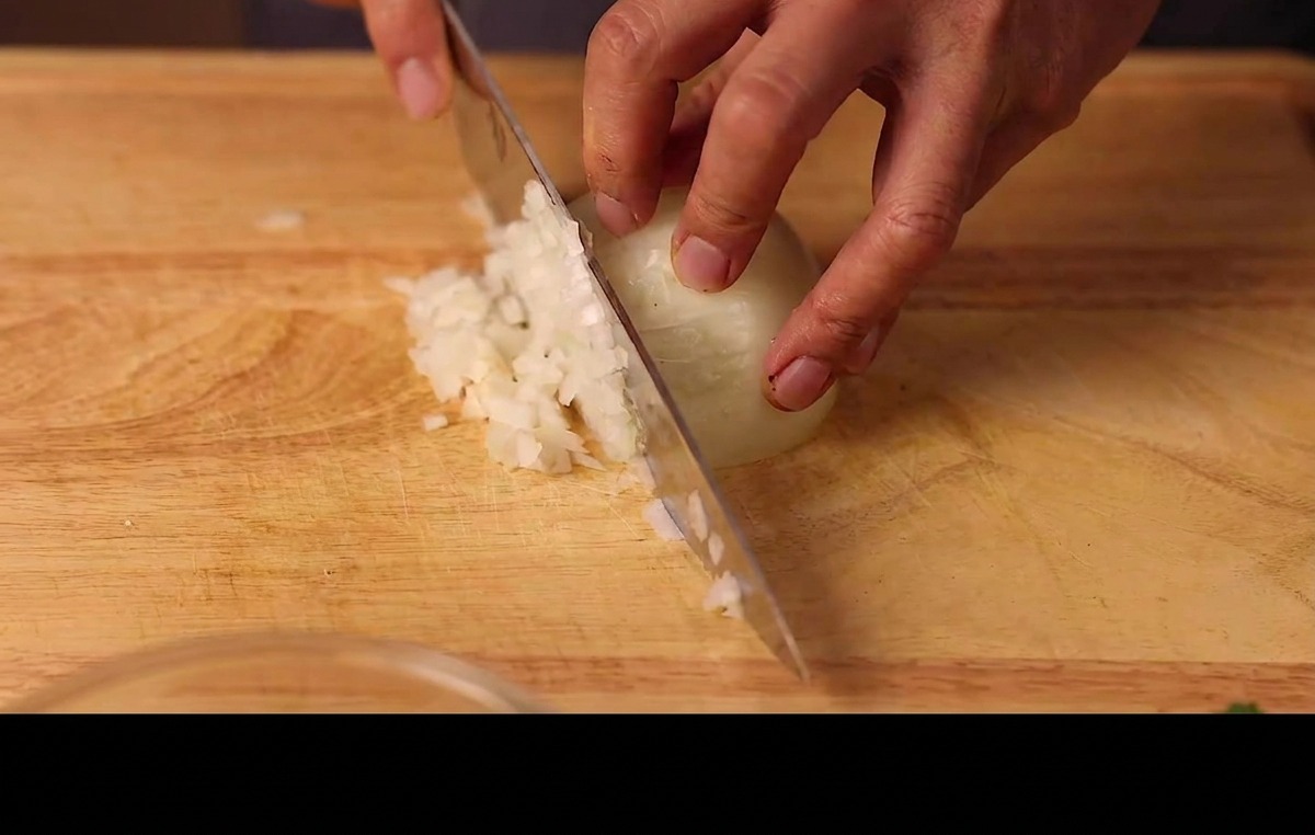 Hands mincing a half of a white onion into fine pieces on a wooden chopping board.