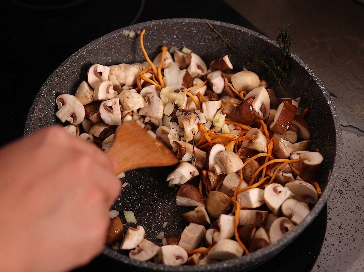 A wooden spatula stirring minced white onions and sliced garlic into a pan of cooked mushrooms.