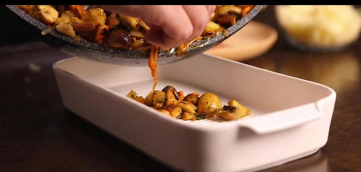 Tipping a frying pan to pour a cooked mixture of chopped mushrooms into a white ceramic baking dish.
