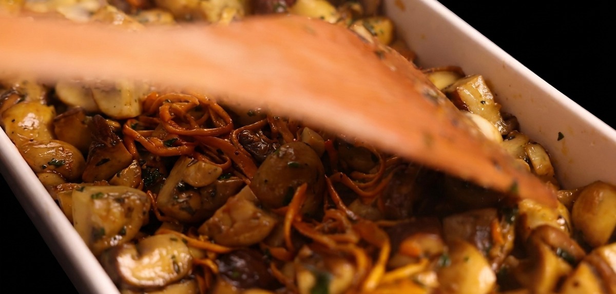 A large wooden spatula flattening a layer of sautéed mushrooms inside a white rectangular baking dish.