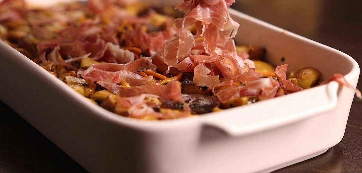 A hand gently dropping curls of thinly sliced ham evenly over a bed of cooked mushrooms in a baking dish.