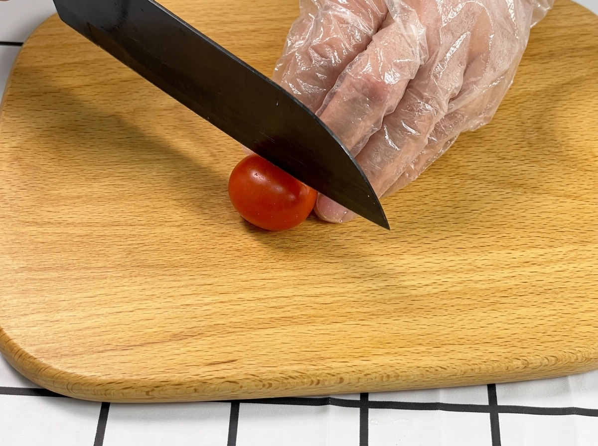 A gloved hand holding a cherry tomato while a black knife slices it in half on a wooden cutting board.