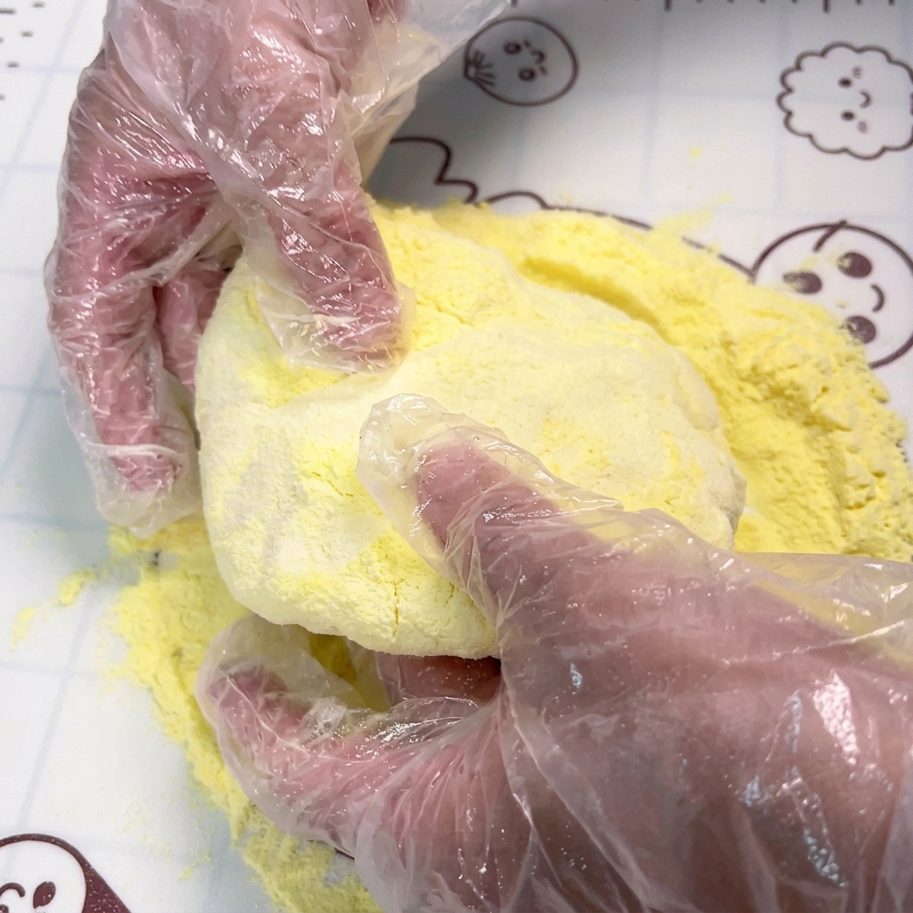 Gloved hands pressing and stretching a round piece of dough over a surface covered in yellow cornstarch.