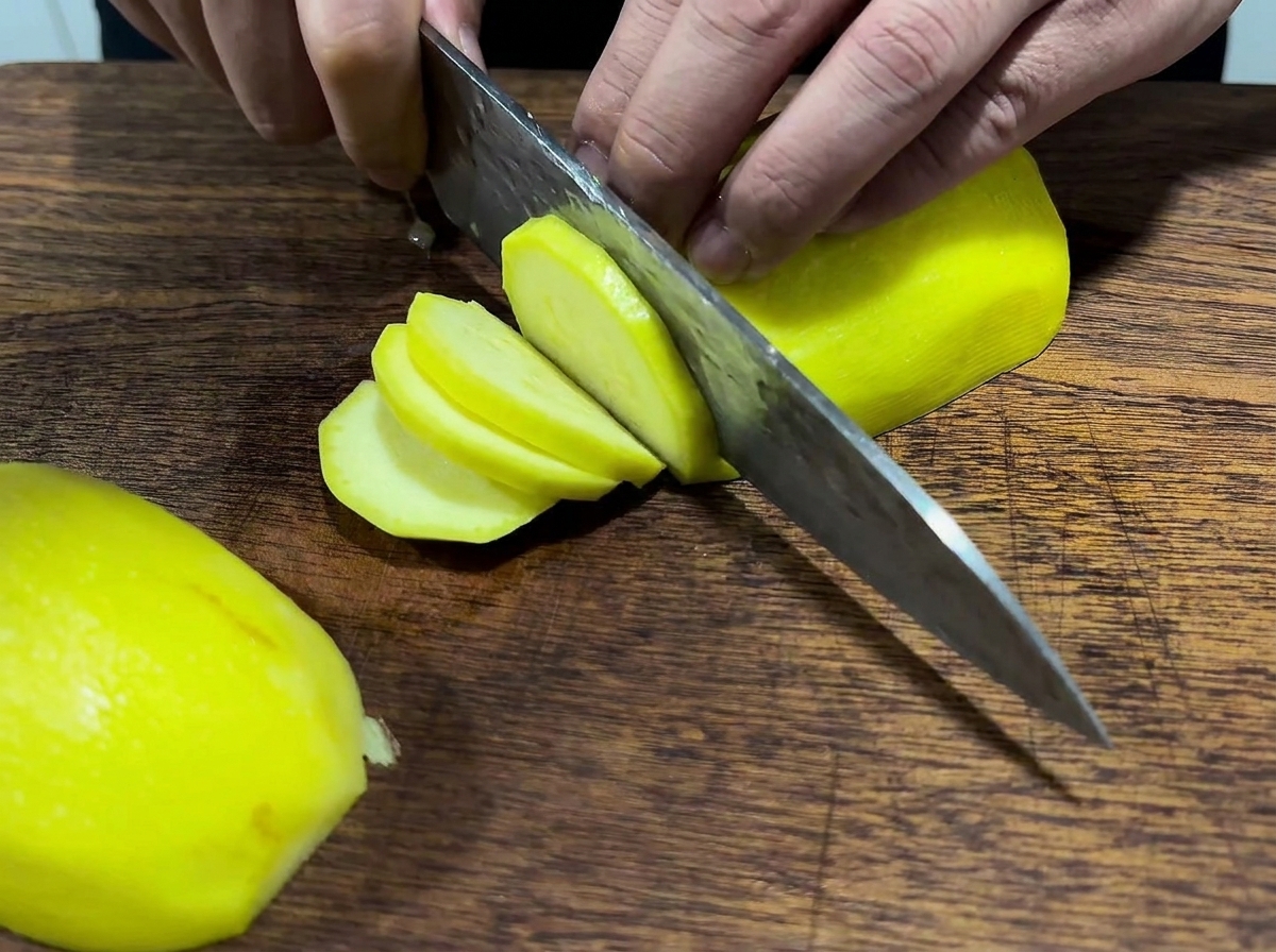 Hands using a large knife to slice a bright yellow peeled potato on a wooden cutting board.
