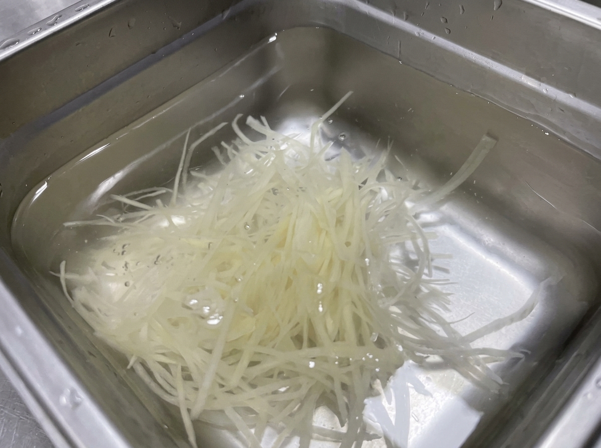 Julienned potato shreds submerged in a stainless steel pan of clear water.