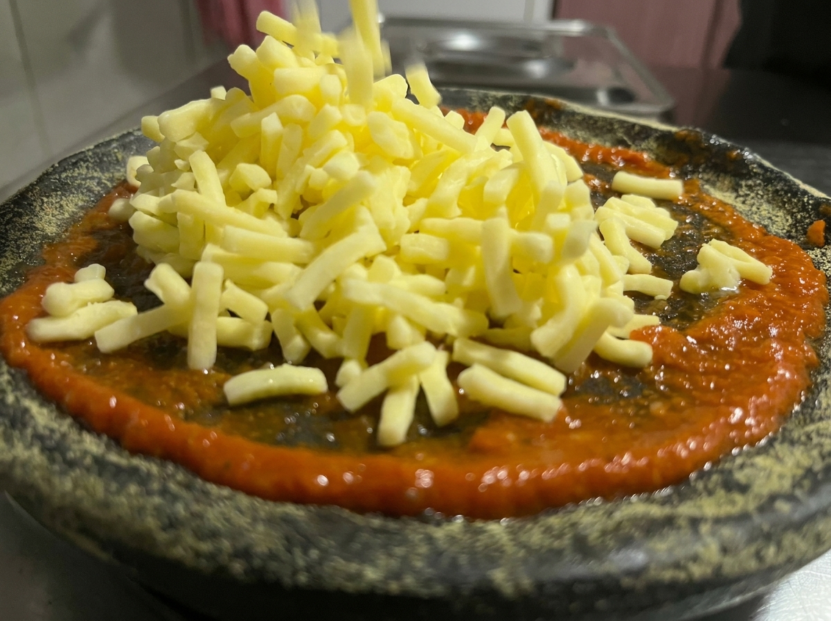A close up of shredded yellow cheese falling onto a layer of bright red tomato sauce spread over a dark charcoal pizza crust.