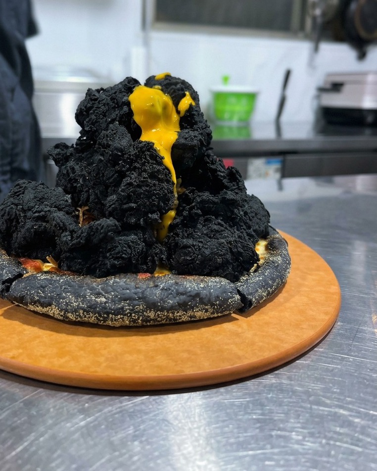 Bright yellow cheese sauce dripping like lava down a tall mound of blackened fried chicken on a black pizza crust.
