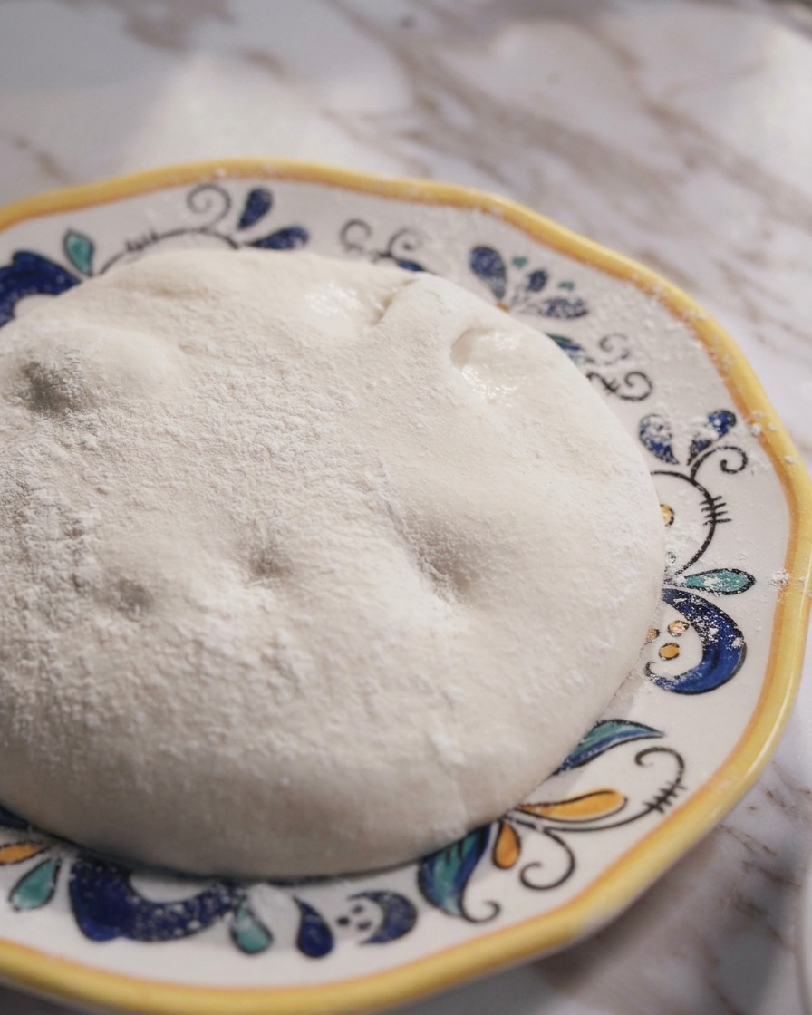 A smooth, round ball of fermented Neapolitan pizza dough sitting on a decorative ceramic plate.