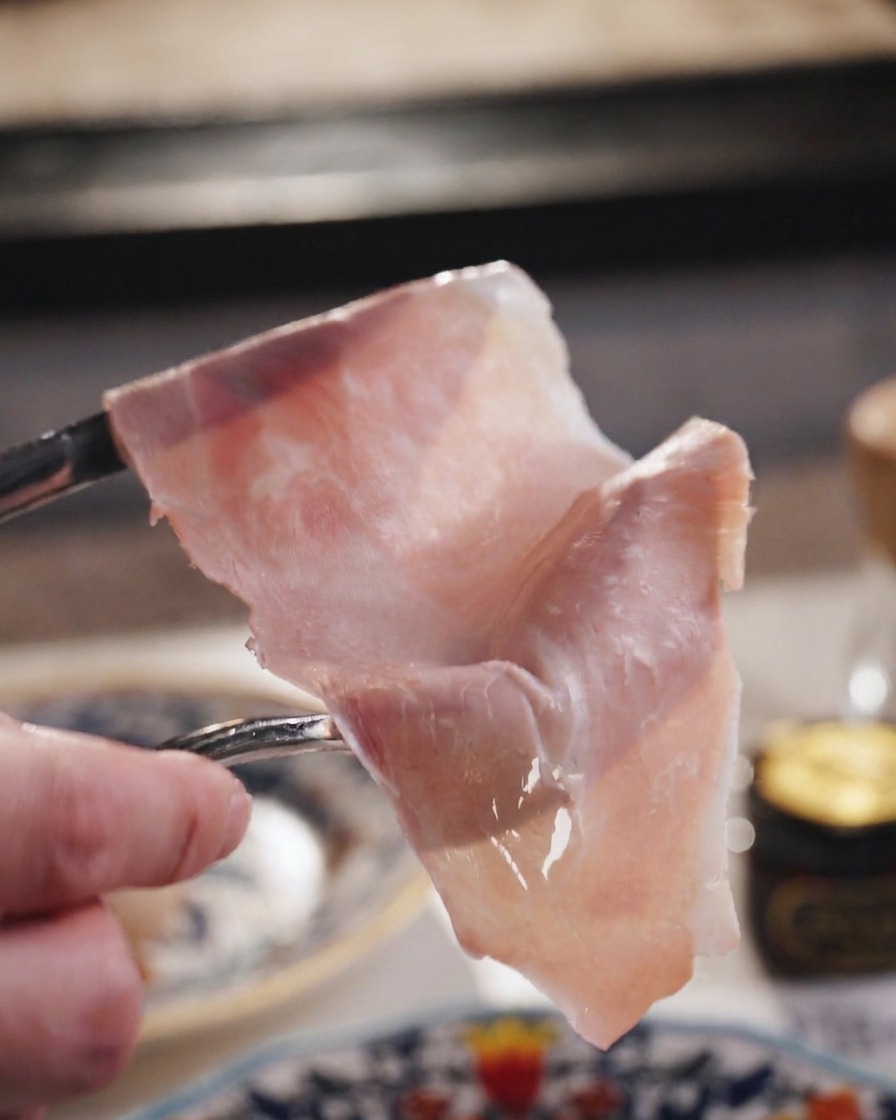 A thin, translucent slice of pink ham held up by a fork against a blurred background.