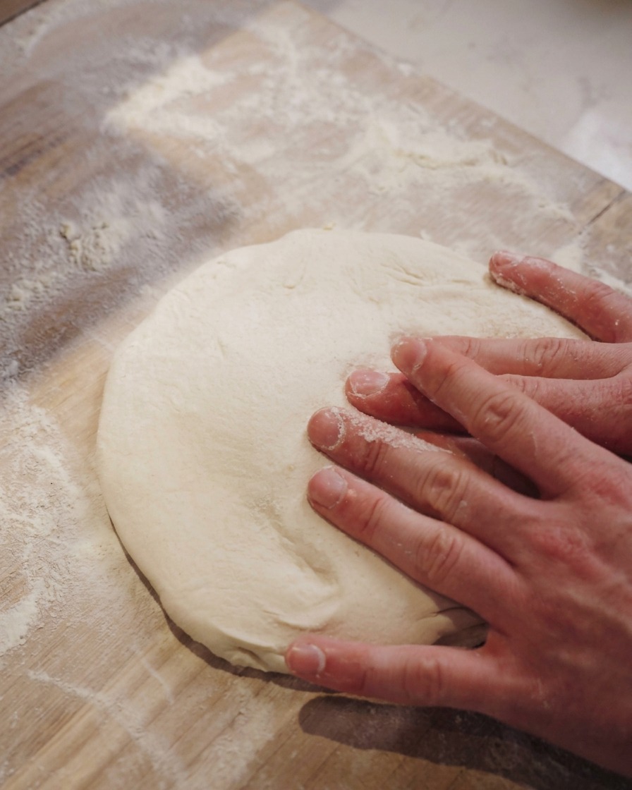 Two hands gently pressing the center of a soft pizza dough round on a wooden surface dusted with flour.