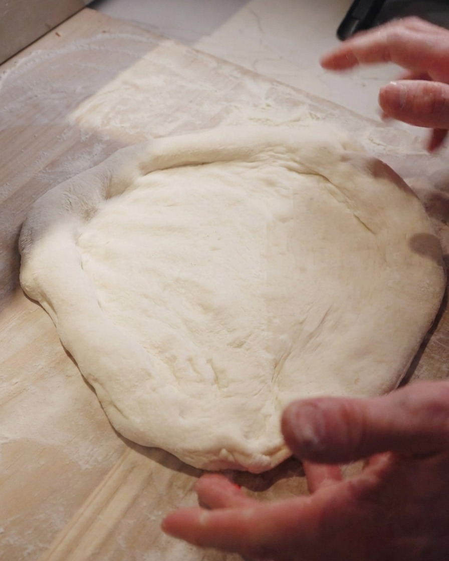 A chefs hands stretching out a pizza dough base on a wooden counter.