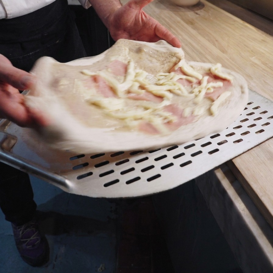 A chef holding a raw pizza on a metal peel, topped with a creamy truffle base, pink ham slices, and strips of white fior di latte cheese.