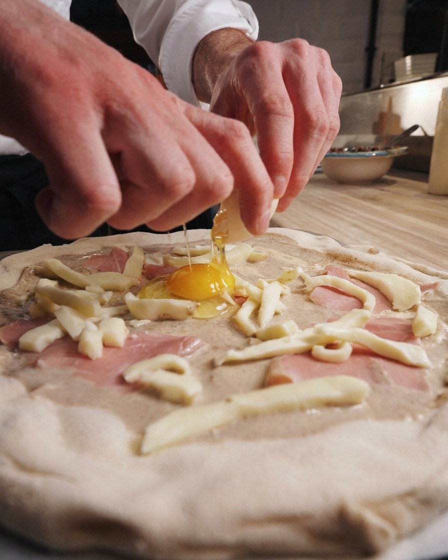 A chefs hands cracking a fresh egg directly onto the center of a raw pizza topped with truffle cream, ham, and fior di latte.