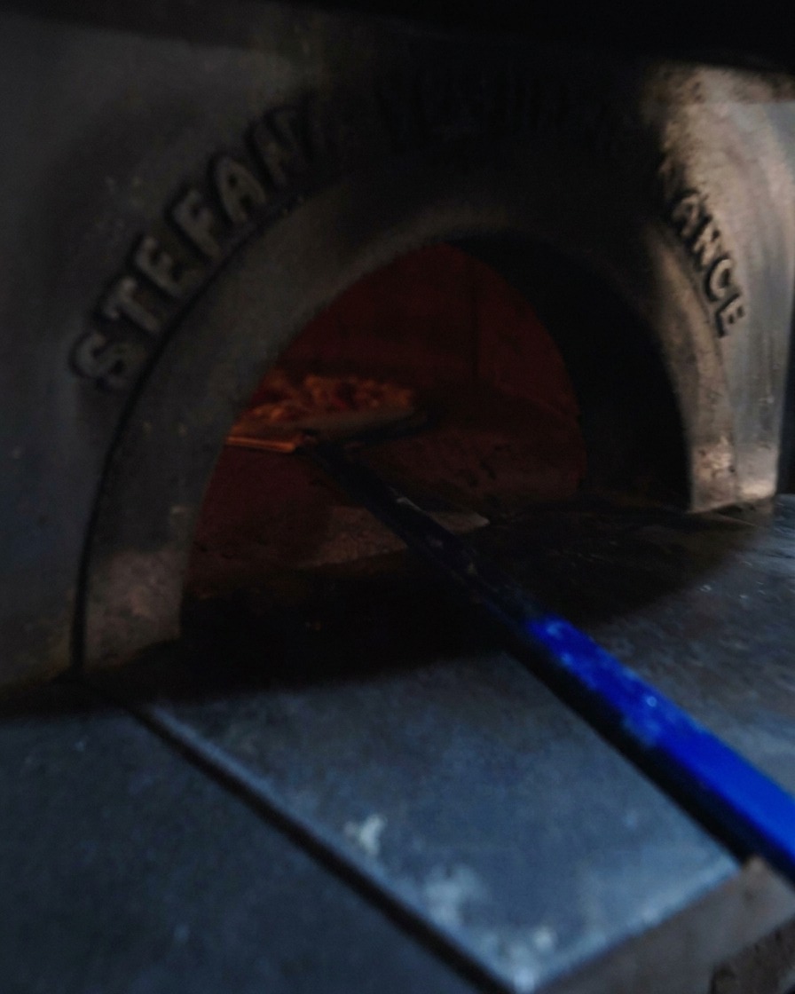 A blue metal pizza peel sliding an assembled Neapolitan pizza into a glowing, hot wood-fired oven.