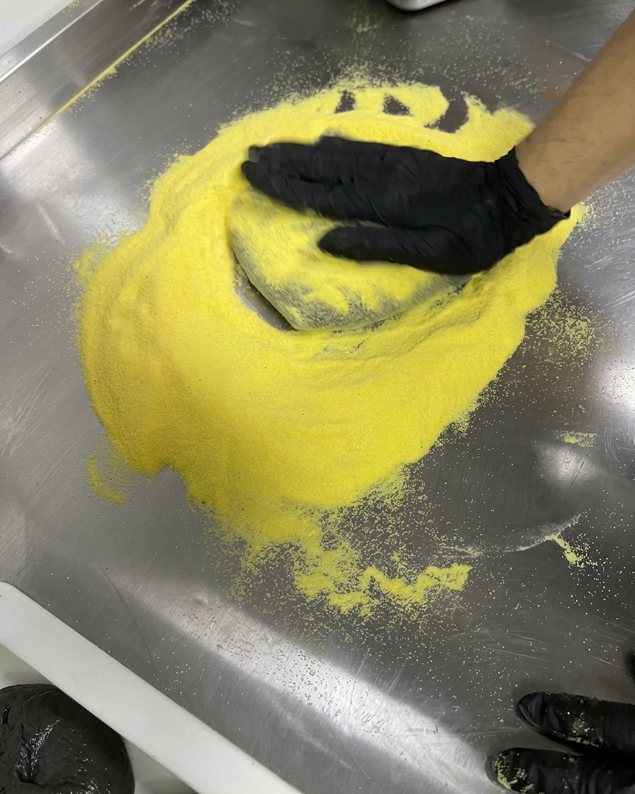 Gloved hands stretching black dough into a circle on a yellow flour dusted steel surface.