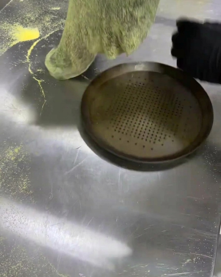 Transferring a flat sheet of black dough onto a perforated metal pizza pan.