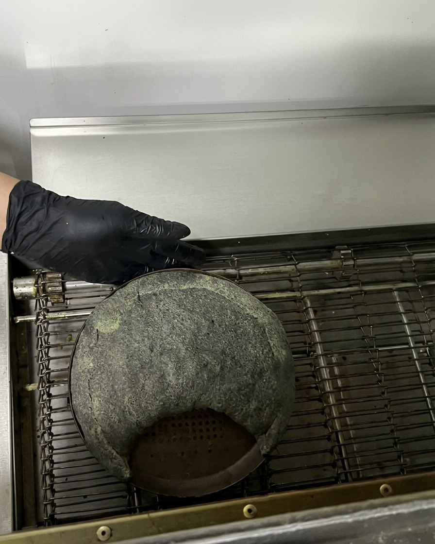 A raw black calzone on a metal pan entering a commercial conveyor belt oven.