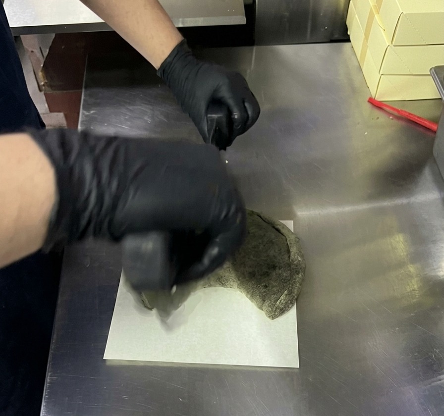 Slicing a baked black calzone with a rocking pizza knife on a white board.