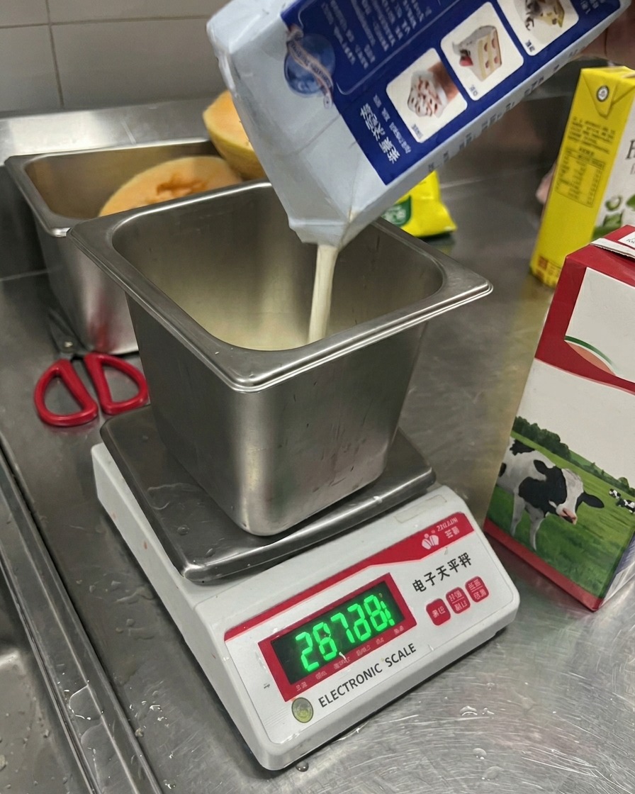 Milk being poured from a carton into a square metal container resting on a digital electronic scale.