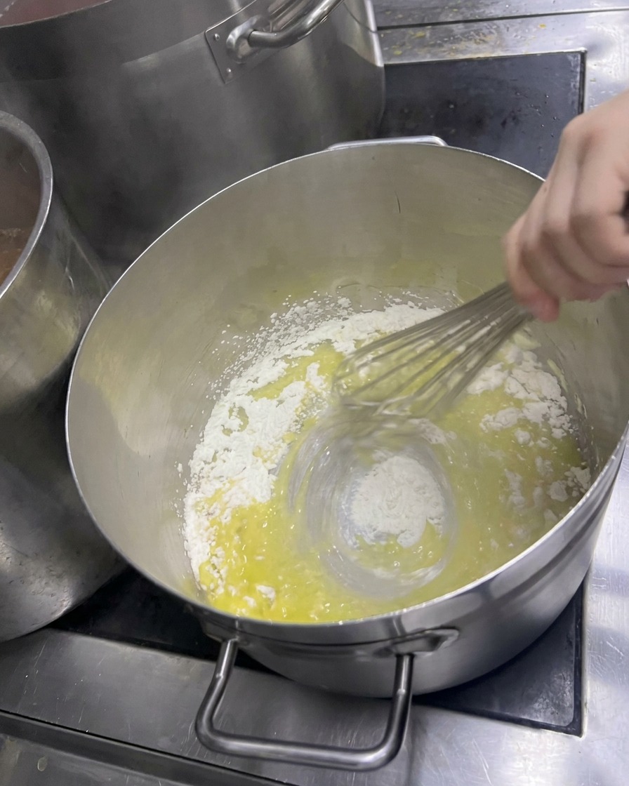 A hand vigorously whisking flour into melted butter in a pot to create a pale yellow roux.