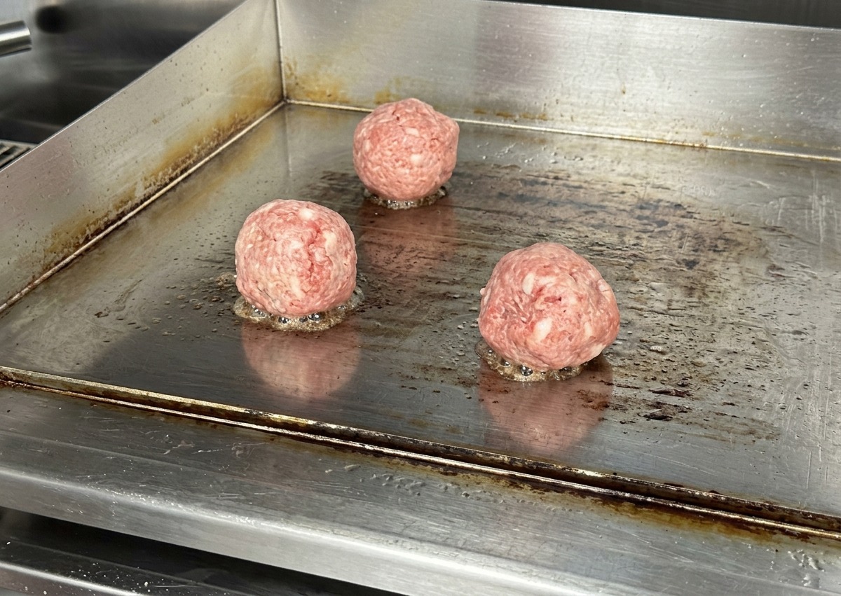 Three round, raw beef balls sitting on a hot, stainless steel flat-top griddle with slight sizzle at the base.