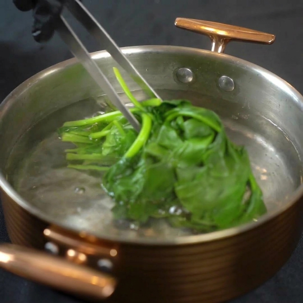Metal tongs lifting bright green blanched spinach leaves out of a pot of boiling water.