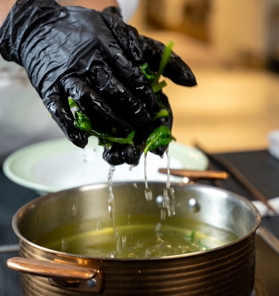 Hands wearing black gloves squeezing water out of a handful of cooked spinach over a pot.