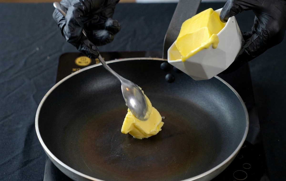 A gloved hand using a spoon to add a block of yellow butter from a white bowl into a heated black frying pan.