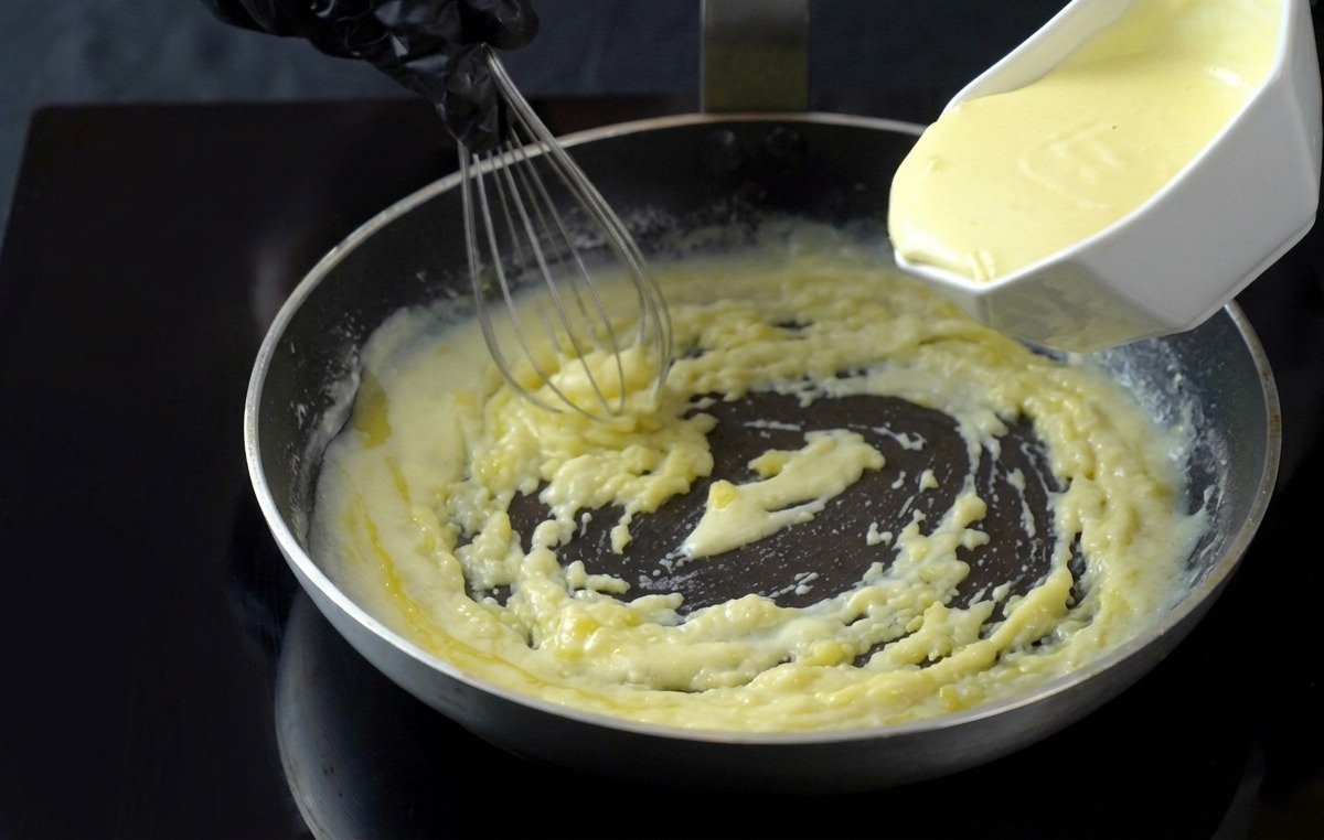 Pouring heavy cream from a white container into a simmering white sauce in a pan, mixing it with a whisk.