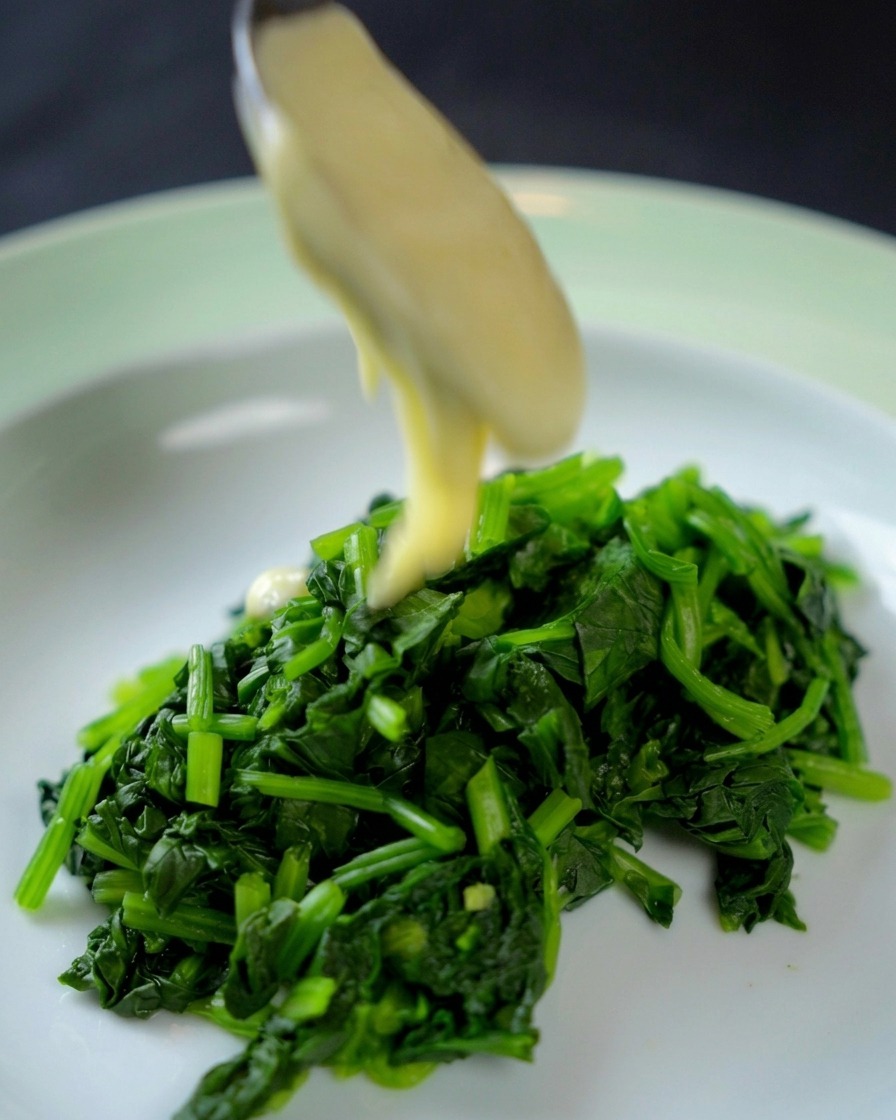 A spoon pouring thick, creamy white sauce over a mound of vibrant, finely chopped spinach in a shallow bowl.