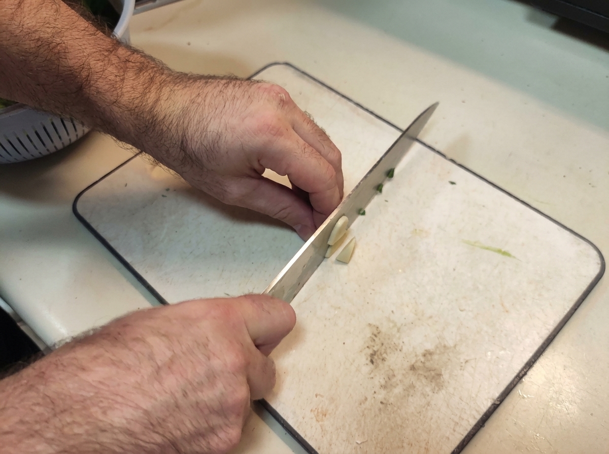 Hands using a chefs knife to finely dice garlic cloves on a white cutting board.