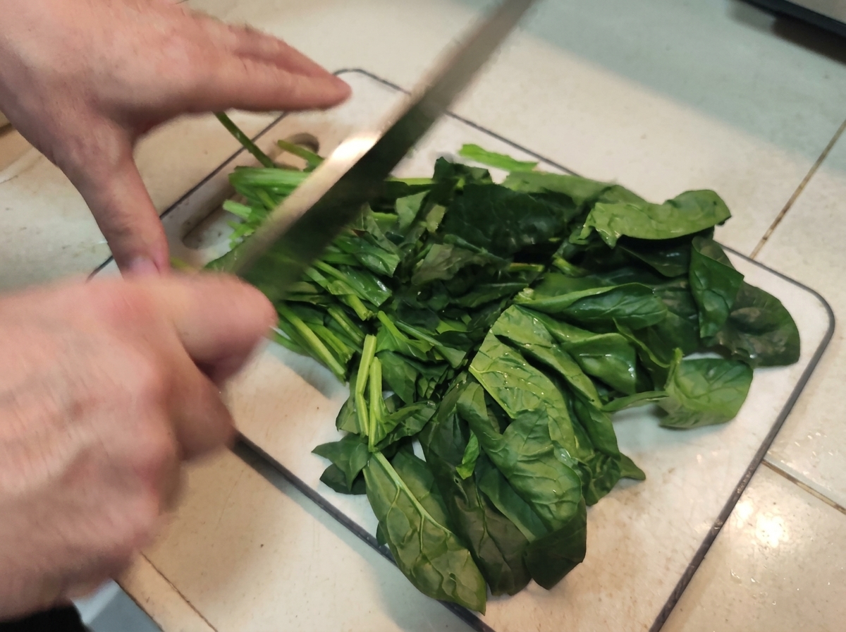 Hands using a large knife to roughly chop a pile of fresh green spinach leaves.