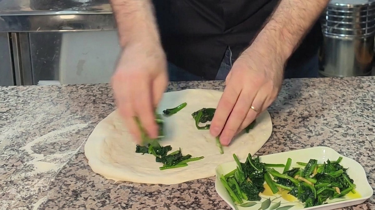 Hands distributing vibrant green cooked spinach evenly over a hand-stretched raw pizza dough base on a floured countertop.