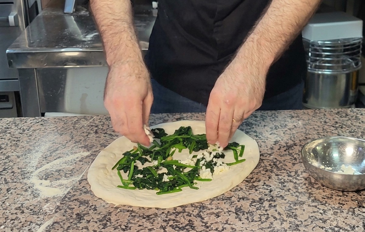 Hands sprinkling crumbled feta cheese over a layer of green spinach on pizza dough.