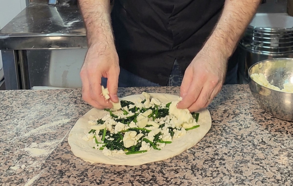 Adding chunks of mozzarella cheese on top of spinach and feta on an unbaked pizza.