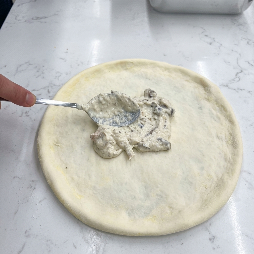 A hand using a metal spoon to spread white creamy mushroom sauce onto a raw pizza dough.
