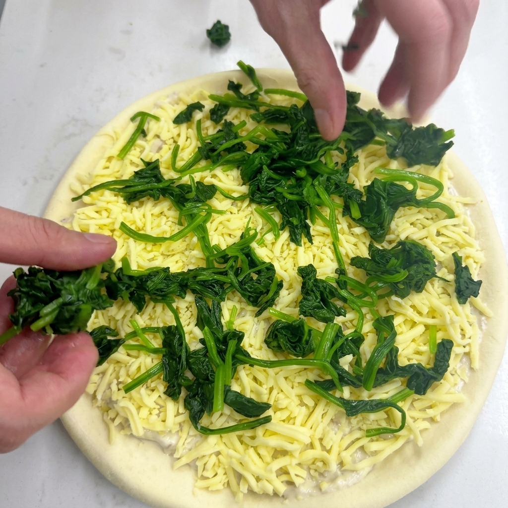 Two hands placing clusters of blanched green spinach onto a pizza base topped with shredded mozzarella cheese.