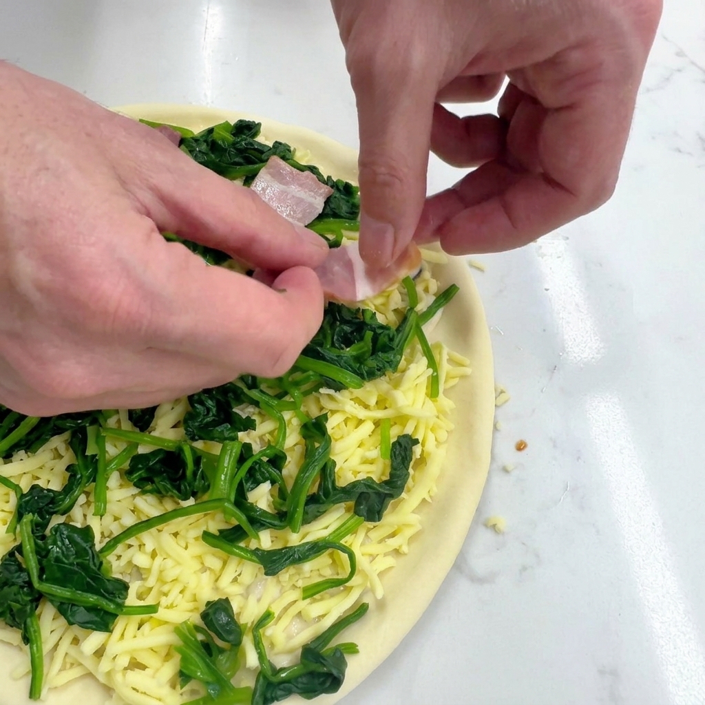Hands carefully placing raw, rectangular bacon slices onto a pizza topped with cheese and green spinach.