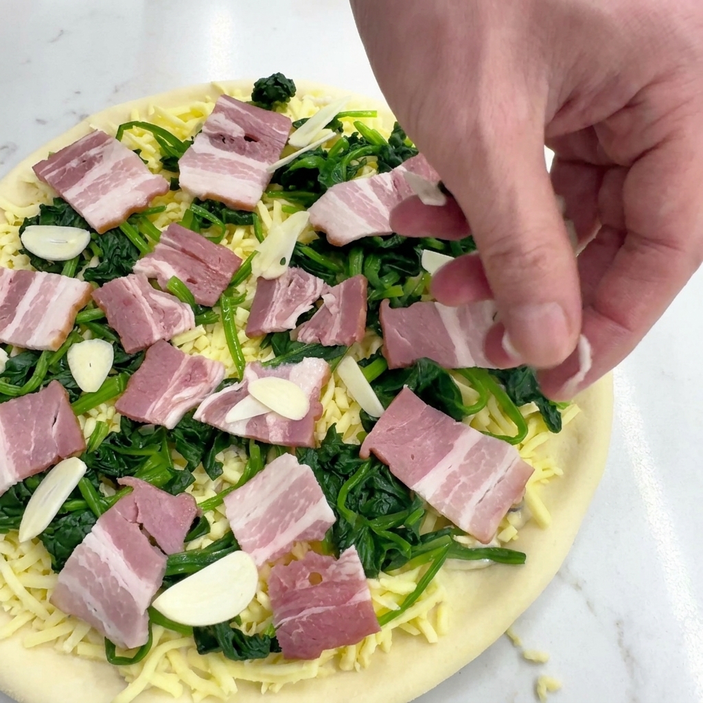 A hand scattering thin slices of raw garlic over a pizza layered with spinach, bacon, and mozzarella.