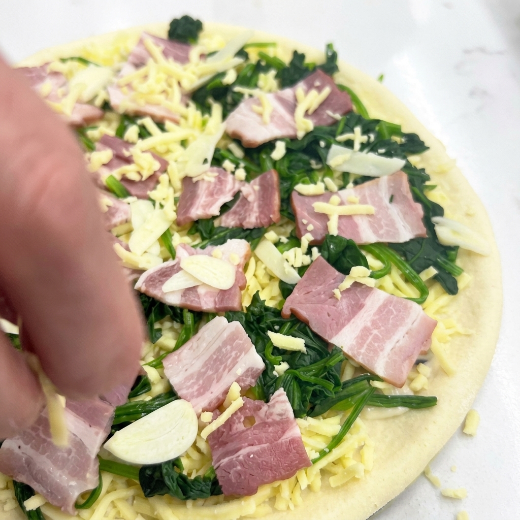 A hand sprinkling shredded mozzarella cheese over an uncooked pizza topped with spinach, bacon, and garlic slices.