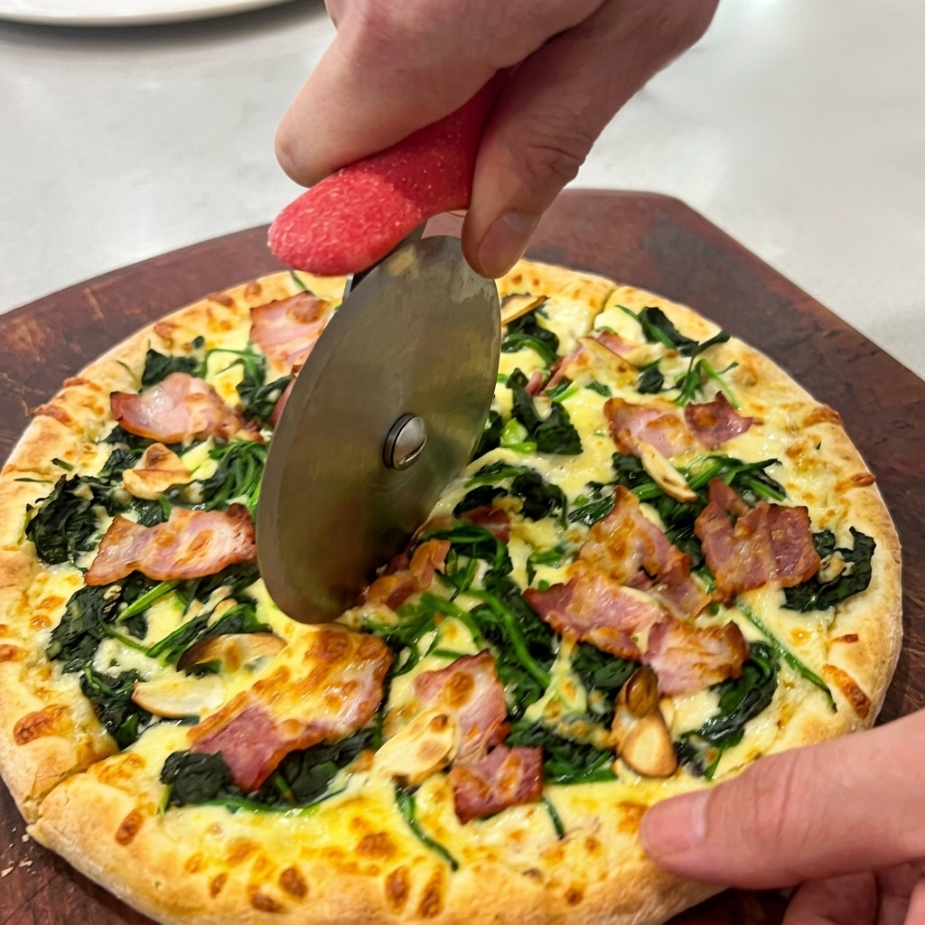 Hands using a circular pizza wheel to slice a hot spinach and bacon pizza on a dark wooden board.