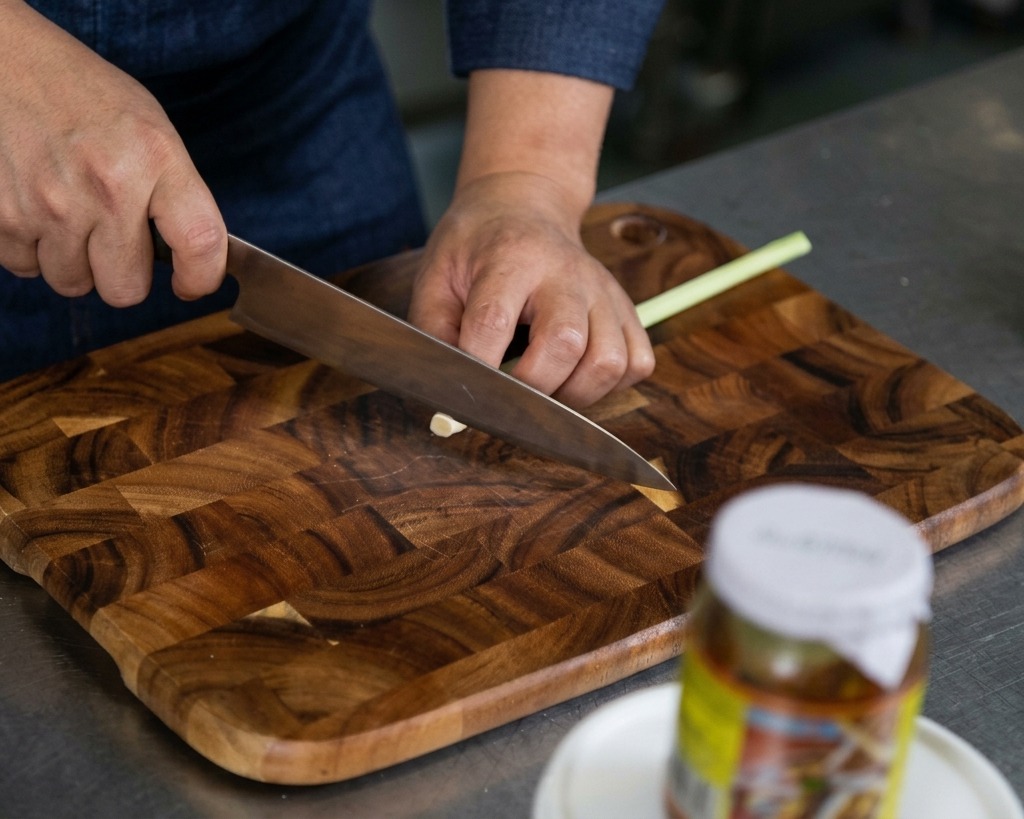 Hands using a sharp knife to finely mince fresh lemongrass stalks on a wooden cutting board.