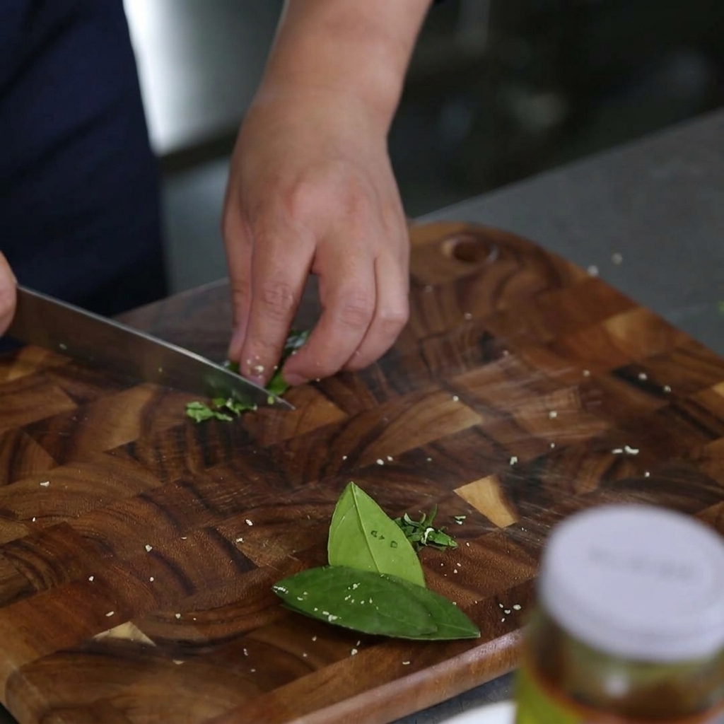 Close up of hands using a chefs knife to slice vibrant green kaffir lime leaves into very thin strips on a wooden cutting board.