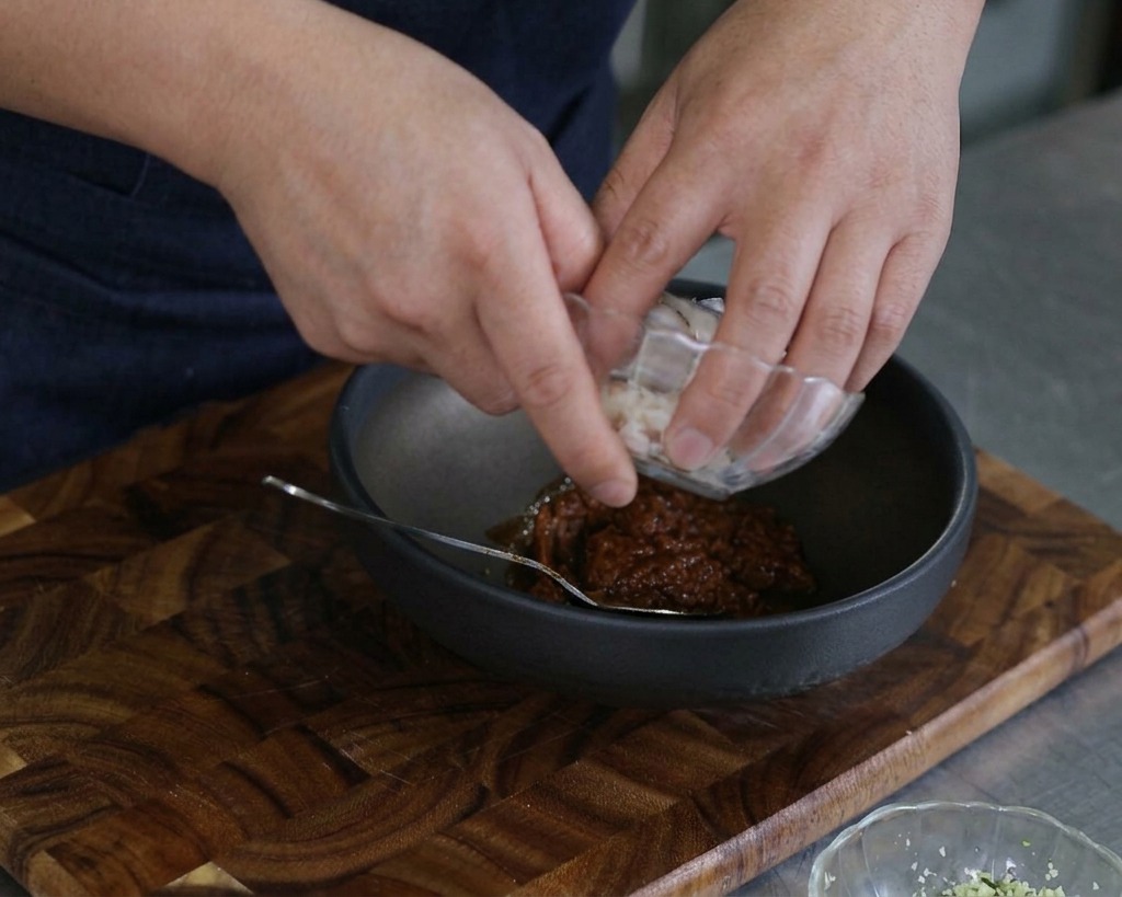 Hands carefully adding raw fish slices and clam meat from a small glass dish into a dark mixing bowl.
