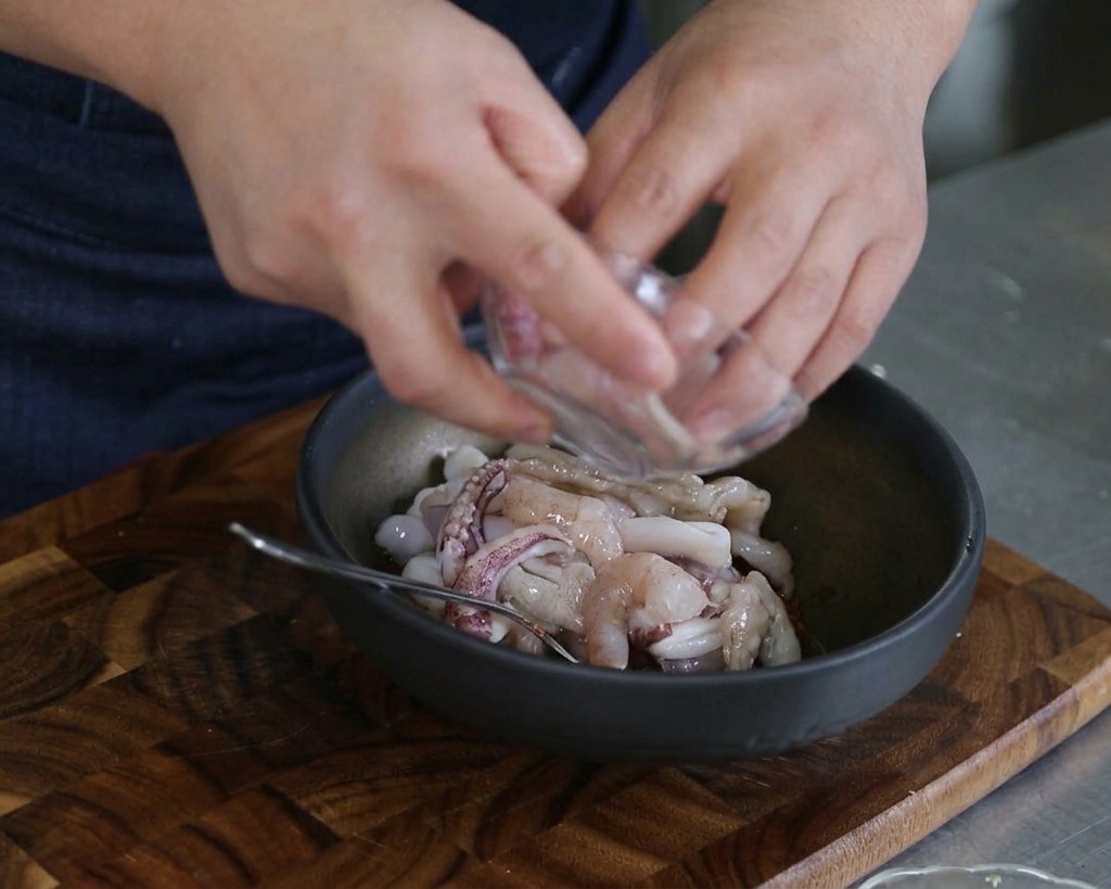 Hands pouring raw sliced squid and shrimp into a dark mixing bowl filled with sauce and other seafood.