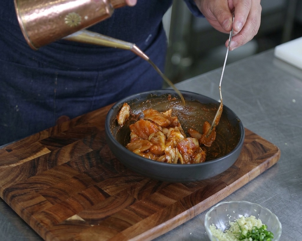 Hands pouring a stream of olive oil from an elegant copper dispenser into a bowl of marinated mixed seafood.